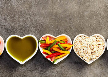 Healthy cooking ingredients placed in long straight row of small heart-shaped bowls, studio shot from above over grey background cropped in wide frame for banner. Organic food concept